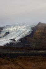 A landscape showcasing a large glacier descending from a mountain, with a blend of icy textures and exposed rocky surfaces. The glacier is partially shrouded in mist, creating a mysterious and serene atmosphere. The foreground consists of a rugged meadow with sparse vegetation.