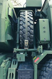 A close-up view of a large military vehicle showcasing the spare tire mounted securely with a strap. The vehicle is predominantly green with various bolts, metal plates, and mechanical components visible. There are red and yellow caution labels affixed to the body.