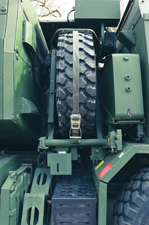 A close-up view of a large military vehicle showcasing the spare tire mounted securely with a strap. The vehicle is predominantly green with various bolts, metal plates, and mechanical components visible. There are red and yellow caution labels affixed to the body.