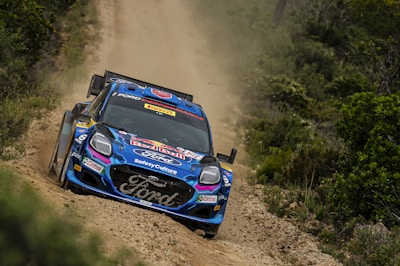 A rally car with various sponsorship logos races along a rugged dirt track surrounded by dense greenery. Dust and debris are stirred up as the car speeds along the uneven path, suggesting a high-speed performance.