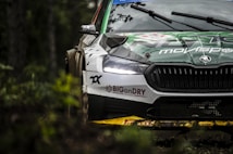 A racing car is navigating through a forested area, with part of the car's design visible, including logos and branding. The focus is on the front end of the car, highlighting its headlights and grille. The surrounding foliage is blurred, suggesting motion and speed.