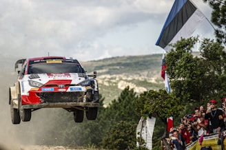 A rally car is airborne, captured in mid-jump during a competitive race on a rugged landscape. Spectators nearby, holding flags and cameras, eagerly watch the vehicle's performance. The setting is outdoors with a partially cloudy sky and green vegetation.