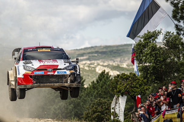A rally car is airborne, captured in mid-jump during a competitive race on a rugged landscape. Spectators nearby, holding flags and cameras, eagerly watch the vehicle's performance. The setting is outdoors with a partially cloudy sky and green vegetation.