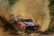The rally car roaring through a muddy river crossing, splashing water everywhere.