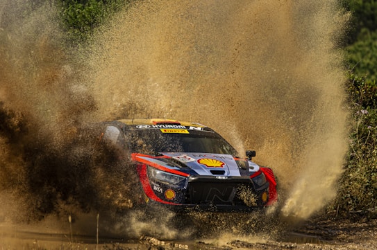 A rally car is speeding through a muddy terrain, splashing water and mud high into the air. The car is adorned with various sponsor logos, including prominent branding from Hyundai and Shell. The vibrant colors of the car contrast with the earthy tones of the mud and surrounding greenery.
