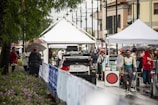 A bustling rally service park featuring race cars and teams preparing for an event. Several individuals are gathered under tents, dressed in racing suits and team jackets. The setting appears lively and organized amidst a light rain, with spectators holding umbrellas and observing the preparations.