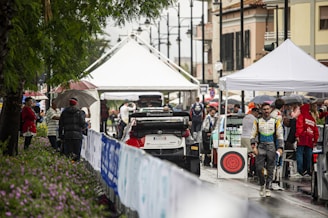 A bustling rally service park featuring race cars and teams preparing for an event. Several individuals are gathered under tents, dressed in racing suits and team jackets. The setting appears lively and organized amidst a light rain, with spectators holding umbrellas and observing the preparations.
