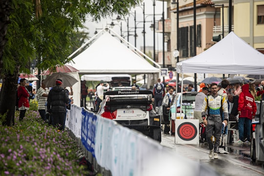 A bustling rally service park featuring race cars and teams preparing for an event. Several individuals are gathered under tents, dressed in racing suits and team jackets. The setting appears lively and organized amidst a light rain, with spectators holding umbrellas and observing the preparations.