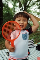 A young child holding a bright orange racket stands in a outdoor setting. The child is wearing a white shirt with a blue pocket and has a cheerful expression. Surrounding them are trees and a striped blanket on the ground.