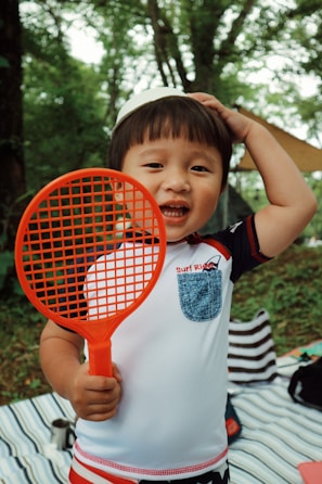 A young child holding a bright orange racket stands in a outdoor setting. The child is wearing a white shirt with a blue pocket and has a cheerful expression. Surrounding them are trees and a striped blanket on the ground.