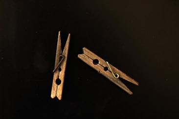 Two wooden clothespins are arranged on a dark background. One is upright while the other is lying horizontally. The wood appears aged, and the metal springs are clearly visible.