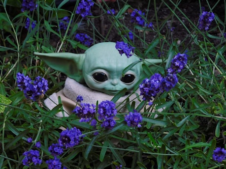 A small green alien-like figure with large ears and big black eyes is nestled among vibrant purple flowers and green foliage.