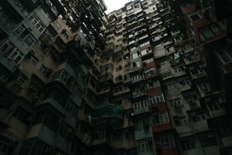 A densely packed residential building with numerous windows and air conditioning units. The architectural style is utilitarian, with a repetitive pattern of balconies and panels reaching up to a small patch of sky. The building's facade is weathered, suggesting age and wear.