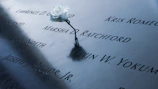 A single white rose placed on a battlefield cross made of a rifle, helmet, and boots.