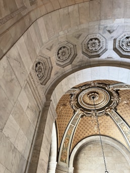 A detailed view of an ornate architectural ceiling with intricate floral and geometric designs. The ceiling features hexagonal and circular patterns with decorative embellishments and a hanging chain from an ornate circular centerpiece. The walls and arches are constructed from a light-colored stone with detailed engravings.