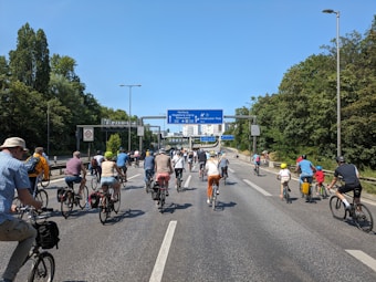 A large group of people riding bicycles on a road that appears to be temporarily closed to vehicular traffic. The cyclists, of varying ages, are moving towards a set of road signs. The environment includes trees on both sides of the road and a clear blue sky.