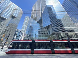 A modern urban scene depicts a sleek red and white streetcar passing in front of towering glass skyscrapers reflecting the blue sky. The architecture showcases contemporary design with grid patterns and reflective surfaces.