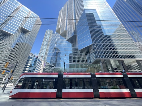A modern urban scene depicts a sleek red and white streetcar passing in front of towering glass skyscrapers reflecting the blue sky. The architecture showcases contemporary design with grid patterns and reflective surfaces.