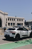 A white autonomous vehicle branded with Waymo is parked on a street next to a large, beige building with arched windows. The sky is clear and blue, and streetlights are visible in the background.