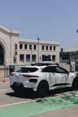 A white autonomous vehicle branded with Waymo is parked on a street next to a large, beige building with arched windows. The sky is clear and blue, and streetlights are visible in the background.