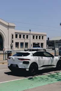 A white autonomous vehicle branded with Waymo is parked on a street next to a large, beige building with arched windows. The sky is clear and blue, and streetlights are visible in the background.