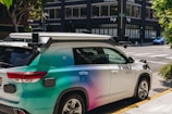 A car equipped with sensors is parked on an urban street corner. The vehicle has the label 'Zoox' on it, indicating it is likely an autonomous vehicle. The car features a futuristic design with various mounted sensors on the roof and sides. The street is lined with modern buildings and there is sparse traffic visible in the background.