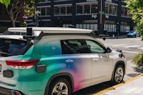 A car equipped with sensors is parked on an urban street corner. The vehicle has the label 'Zoox' on it, indicating it is likely an autonomous vehicle. The car features a futuristic design with various mounted sensors on the roof and sides. The street is lined with modern buildings and there is sparse traffic visible in the background.