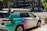 A car equipped with sensors is parked on an urban street corner. The vehicle has the label 'Zoox' on it, indicating it is likely an autonomous vehicle. The car features a futuristic design with various mounted sensors on the roof and sides. The street is lined with modern buildings and there is sparse traffic visible in the background.
