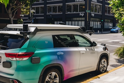 A car equipped with sensors is parked on an urban street corner. The vehicle has the label 'Zoox' on it, indicating it is likely an autonomous vehicle. The car features a futuristic design with various mounted sensors on the roof and sides. The street is lined with modern buildings and there is sparse traffic visible in the background.