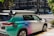A car equipped with sensors is parked on an urban street corner. The vehicle has the label 'Zoox' on it, indicating it is likely an autonomous vehicle. The car features a futuristic design with various mounted sensors on the roof and sides. The street is lined with modern buildings and there is sparse traffic visible in the background.