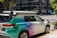 A car equipped with sensors is parked on an urban street corner. The vehicle has the label 'Zoox' on it, indicating it is likely an autonomous vehicle. The car features a futuristic design with various mounted sensors on the roof and sides. The street is lined with modern buildings and there is sparse traffic visible in the background.