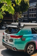 A turquoise autonomous vehicle with multiple sensors is parked on an urban street. It is surrounded by trees and modern buildings, with visible license plates and sleek design details.