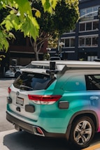 A turquoise autonomous vehicle with multiple sensors is parked on an urban street. It is surrounded by trees and modern buildings, with visible license plates and sleek design details.