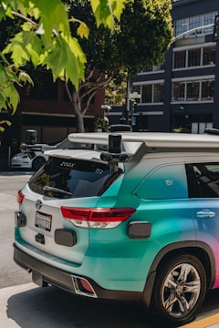 A turquoise autonomous vehicle with multiple sensors is parked on an urban street. It is surrounded by trees and modern buildings, with visible license plates and sleek design details.