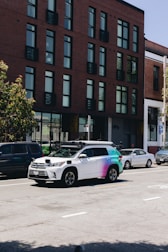 A street scene featuring a row of parked cars along the side of the road with a distinctive white and multicolored SUV equipped with sensors on top, likely a self-driving vehicle. The background shows a modern brick building with large windows.
