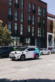 A street scene featuring a row of parked cars along the side of the road with a distinctive white and multicolored SUV equipped with sensors on top, likely a self-driving vehicle. The background shows a modern brick building with large windows.
