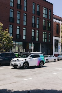 A street scene featuring a row of parked cars along the side of the road with a distinctive white and multicolored SUV equipped with sensors on top, likely a self-driving vehicle. The background shows a modern brick building with large windows.
