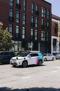 A street scene featuring a row of parked cars along the side of the road with a distinctive white and multicolored SUV equipped with sensors on top, likely a self-driving vehicle. The background shows a modern brick building with large windows.
