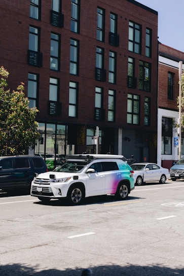 A street scene featuring a row of parked cars along the side of the road with a distinctive white and multicolored SUV equipped with sensors on top, likely a self-driving vehicle. The background shows a modern brick building with large windows.