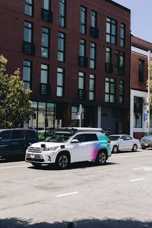 A street scene featuring a row of parked cars along the side of the road with a distinctive white and multicolored SUV equipped with sensors on top, likely a self-driving vehicle. The background shows a modern brick building with large windows.