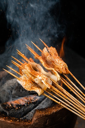 Skewers of food are cooking over a charcoal grill, with smoke and flames rising around them. The skewers appear to hold seasoned slices of meat or tofu, sprinkled with spices. The background is dark, highlighting the warm colors of the food and the orange glow of the embers.