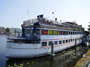 An old, weathered boat with multiple decks is moored on a calm body of water. The boat exhibits signs of rust and wear, with colorful windows in red, yellow, and green. There are decorative lights strung across the top deck, and some plants are visible on the upper level. The surrounding area includes additional boats and modern buildings in the background.