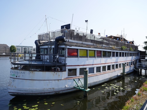 An old, weathered boat with multiple decks is moored on a calm body of water. The boat exhibits signs of rust and wear, with colorful windows in red, yellow, and green. There are decorative lights strung across the top deck, and some plants are visible on the upper level. The surrounding area includes additional boats and modern buildings in the background.