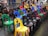 A collection of colorful plastic chairs arranged in rows indoors, each equipped with a set of headphones. The chairs are in various colors, including yellow, green, blue, black, and red. In the background, several people are walking or standing, some carrying bags or wearing casual attire, indicating a public event or gathering.