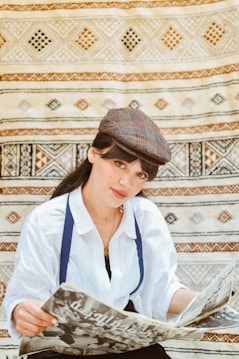 A person wearing a patterned flat cap and a white shirt sits in front of a detailed woven rug or tapestry. They hold a newspaper with Arabic text, appearing relaxed and slightly smiling.