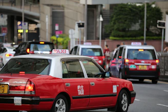 A city street scene featuring several red taxis, each with a white top and a sign indicating they are taxis. The taxis are driving on a multi-lane road, surrounded by other vehicles such as cars and possibly buses. Buildings and greenery can be seen in the background, suggesting an urban environment.