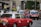 A city street scene featuring several red taxis, each with a white top and a sign indicating they are taxis. The taxis are driving on a multi-lane road, surrounded by other vehicles such as cars and possibly buses. Buildings and greenery can be seen in the background, suggesting an urban environment.