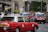 A city street scene featuring several red taxis, each with a white top and a sign indicating they are taxis. The taxis are driving on a multi-lane road, surrounded by other vehicles such as cars and possibly buses. Buildings and greenery can be seen in the background, suggesting an urban environment.