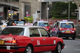 A city street scene featuring several red taxis, each with a white top and a sign indicating they are taxis. The taxis are driving on a multi-lane road, surrounded by other vehicles such as cars and possibly buses. Buildings and greenery can be seen in the background, suggesting an urban environment.