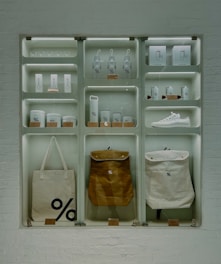A well-organized display of branded merchandise with a minimalist aesthetic. The items include bottles, cups, a sneaker, bags, and other accessories, all featuring a distinctive percentage symbol on a white or neutral background. The display is set against a white brick wall, giving an industrial feel.
